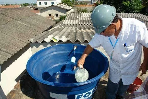 Homem de capacete e camisa branca coleta água de caixa d'água azul em telhado