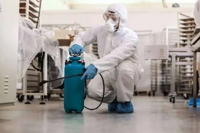 Pessoa com EPI branco e máscara, agachada, usando pulverizador azul para desinfecção em ambiente industrial