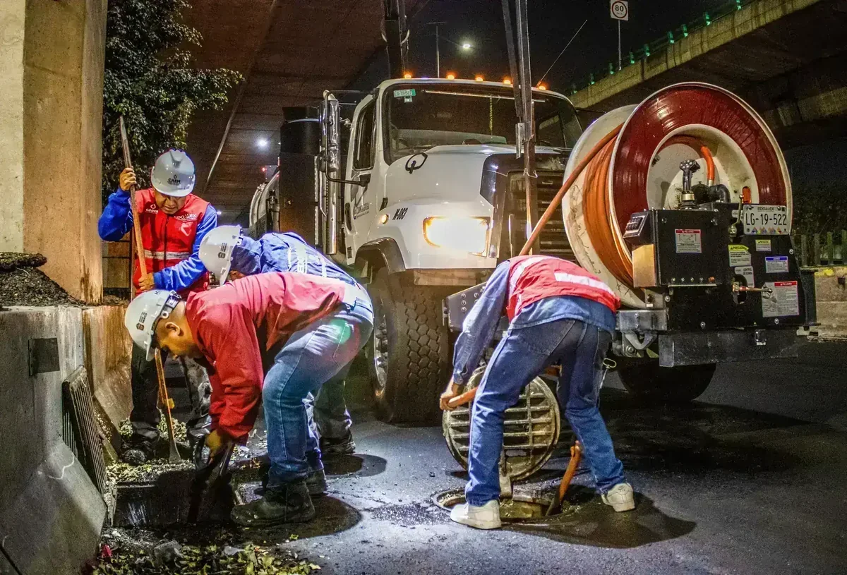 Trabalhadores limpando bueiros e ralos na rua à noite, com caminhão de desentupimento